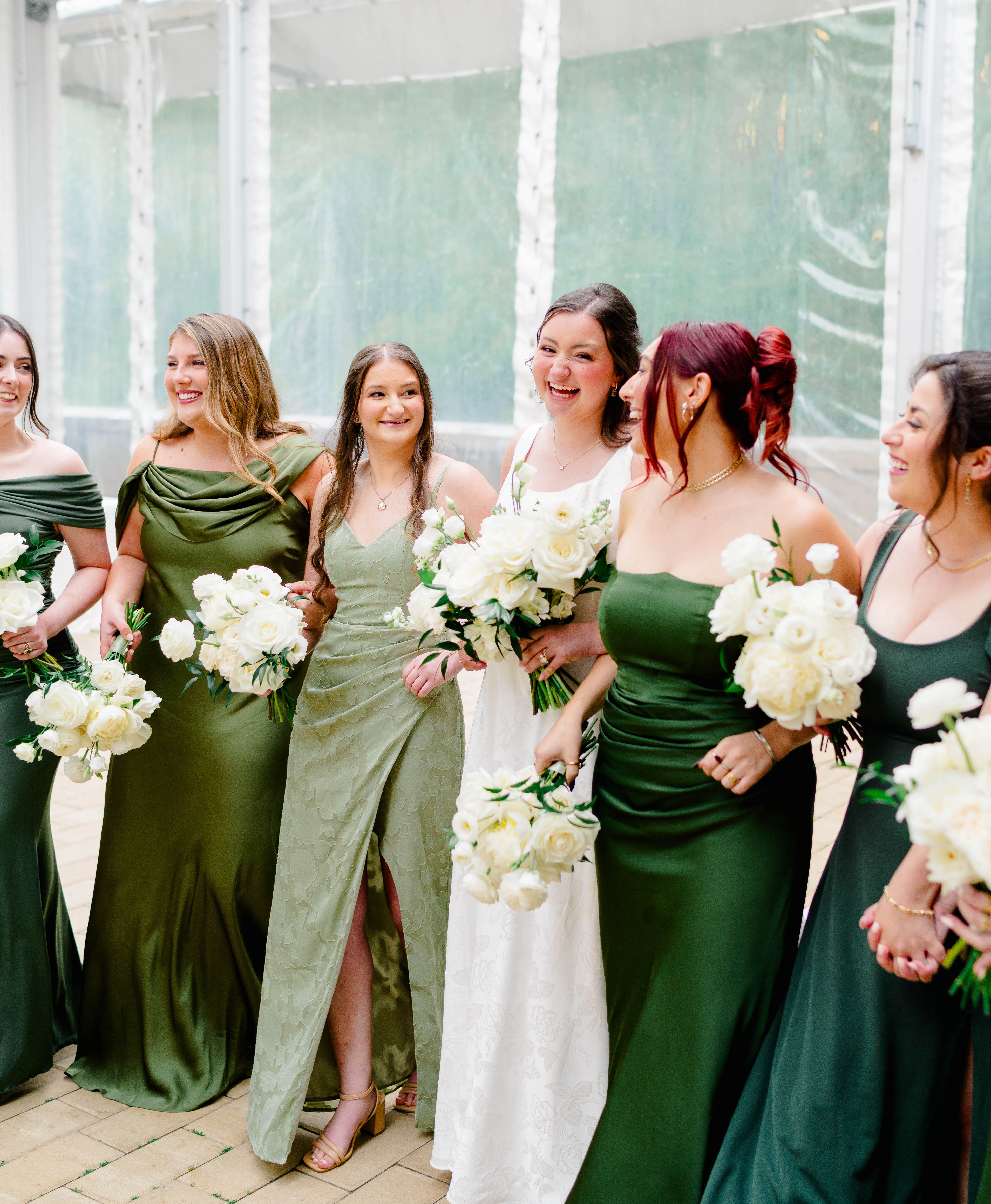 Bride laughing with her bridesmaids in emerald and sage green gowns, all holding white rose bouquets