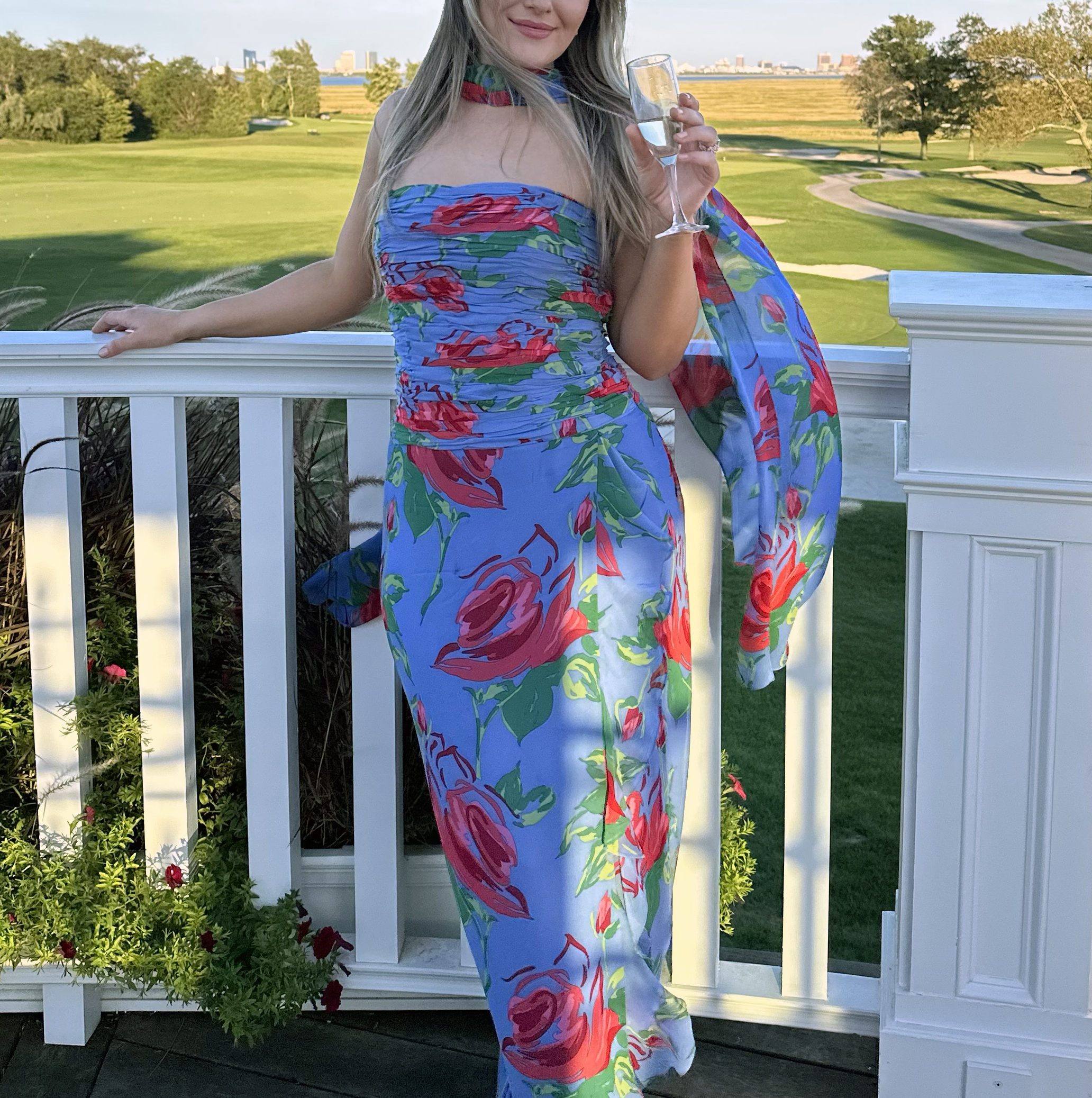 Wedding guest in a blue floral gown with a matching wrap, holding a glass of champagne on a country club terrace